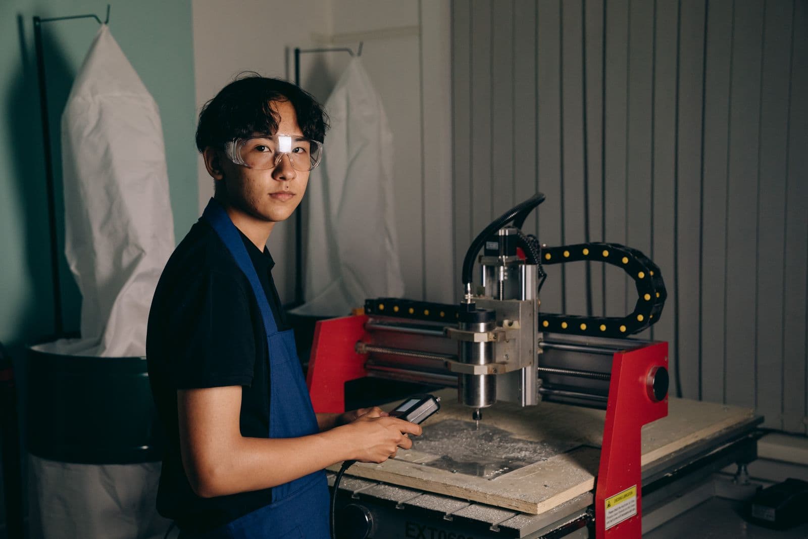 Learner in safety glasses running a benchtop milling machine from its handheld controller during supervised practice.