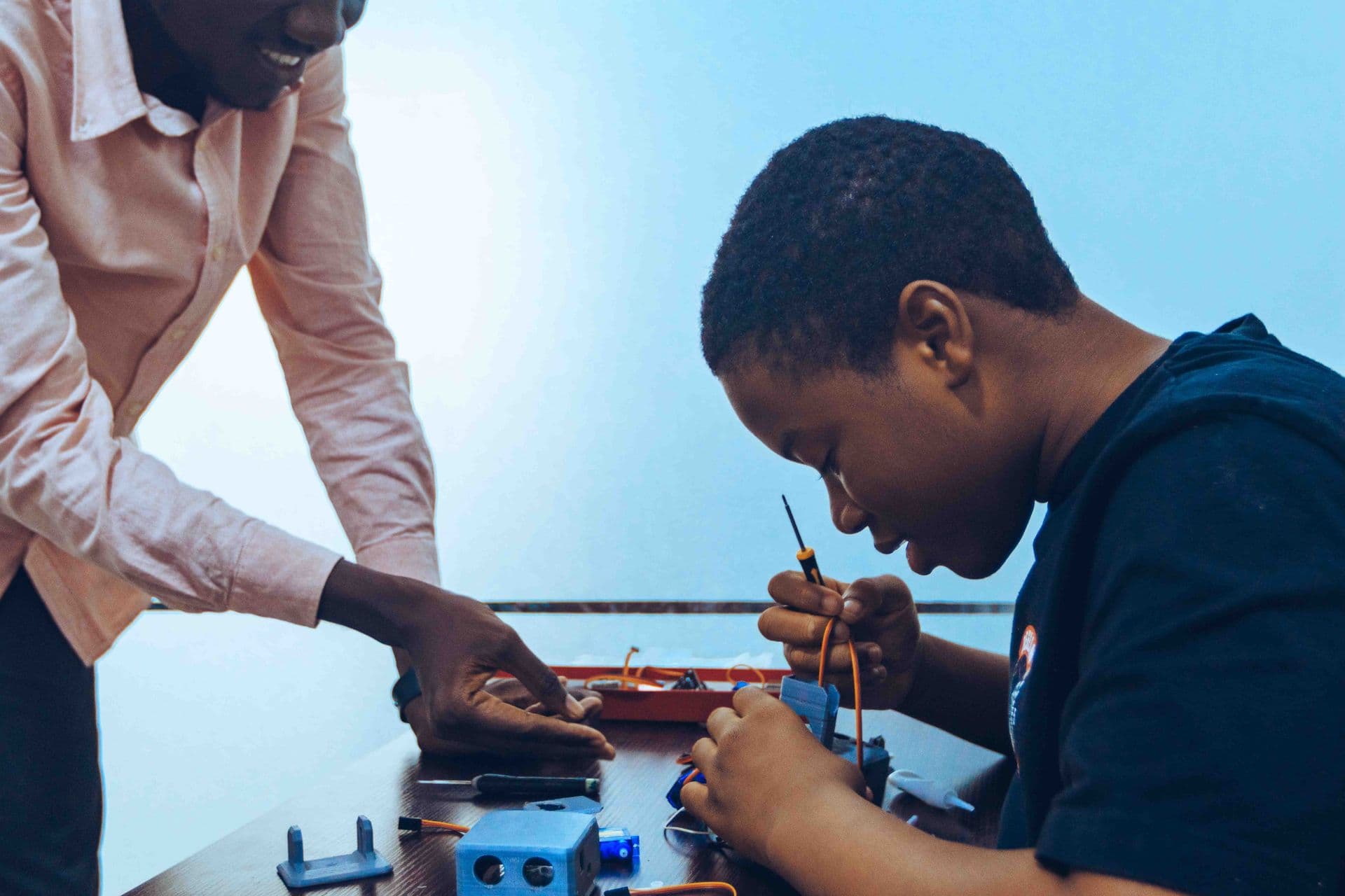 Mentor leaning in to guide a young learner assembling components at a workbench.