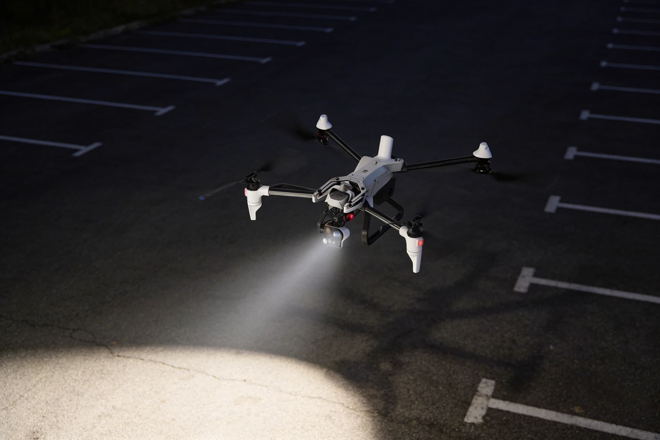 A response drone hovering at night, its spotlight beam sweeping an empty parking lot below